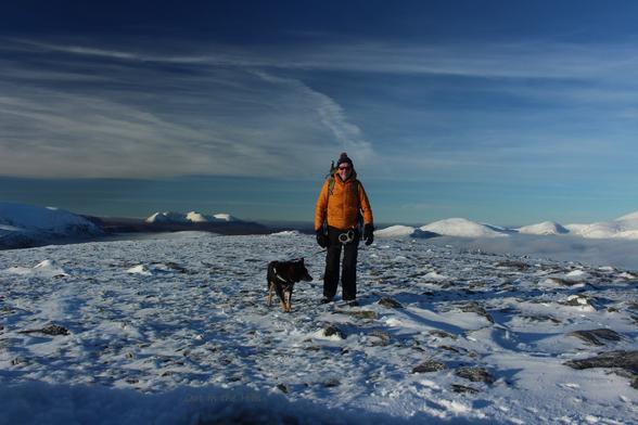 On top of a hill, the ground is rocky but covered in snow. There are snow covered hills in the distance. Blue sky with wispy clouds. Moray the dog and I are stood facing the camera, I am wrapped up well against the cold.