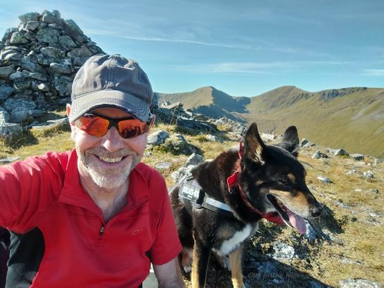 Moray the dog and me sat just below a summit cairn of stones, on a grassy and rocky hill. I am wearing a red short sleeved shirt, baseball cap, and sunglasses. There is a ridge of hills at the back. Blue sky with wispy clouds.