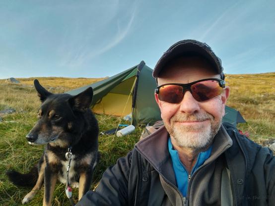 A one person tent on a grassy slope, Moray the dog and I are sat in front of the tent, smiling because there are no midges.