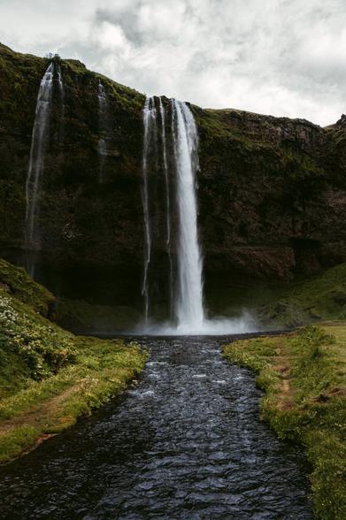The image shows a stunning natural scene of a tall waterfall cascading down a rocky cliff covered in patches of green vegetation. The water falls into a calm stream that flows toward the viewer, framed by grassy banks dotted with wildflowers. The sky above is cloudy, creating a dramatic and slightly moody atmosphere. The entire scene feels serene and untouched, with a balance of rugged nature and gentle greenery. Taken in Iceland by European elopement photography Sturmsucht.