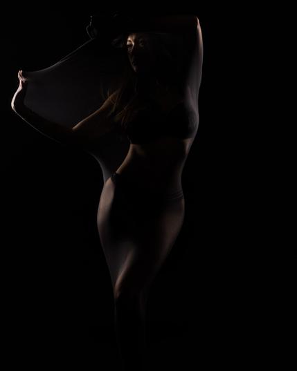 Eine kreative und künstlerische Darstellung einer Frau in einem Nylon-Schauch, die von hinten mit Licht angestrahlt wird und sich in einer dramatischen Pose rekelt.  Man sieht fast nur die Figur/silhouette der Frau.