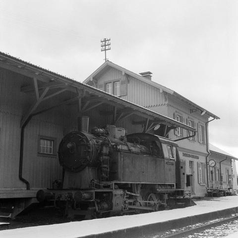 The black and white photograph depicts a steam locomotive parked on a snow-covered railway platform. The locomotive is positioned in the foreground, its large, cylindrical boiler and smokestack dominating the image. The engine is partially obscured by a wooden, roofed platform that extends from the side of a wooden station building. The station building is two stories tall and has several windows. A signal mast stands tall in the background, its crossbar visible against the gray sky. The overall atmosphere of the image is one of tranquility and nostalgia, with the snow-covered platform and the steam locomotive evoking a bygone era of railway travel.