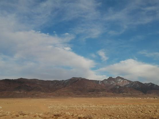 A panoramic view of a dry desert landscape featuring rugged mountains in the background. The mountains are partially covered in snow, while the foreground shows a flat terrain with sparse vegetation. The sky is a mix of blue and white clouds, indicating a clear day.