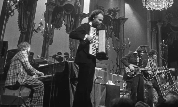 A tall-foreheaded young man in a suit stands on an ornate stage, solemnly playing a piano accordion in a rock ensemble.