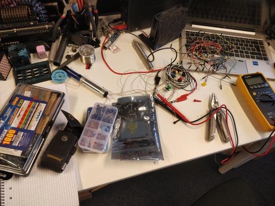 Ett foto av ett skrivbord. På skrivbordet finns en dator, en multimeter, ett multiverktyg, massor av kablar, lödverktyg, elkomponenter i påsar och lådor, 3D-utskrivna prylar, m.m. i en allsköns röra.
