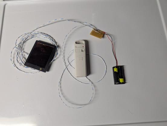 A picture of a plastic temperature sensor, about five inches long and one inch wide, a transmitter used for an indoor/outdoor digital thermometer.  A few feet of twisted pair wire runs out the side to a small prototype board, to which a battery holder with two AA batteries is also connected, along with a small black solar panel connected by about six feet of wire, which is coiled up.  Everything is sitting on a white surface, actually the top of a clothes dryer.