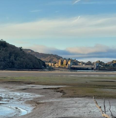 Conwy Castle taken at a distance from across Colwyn Bay. The tide is out, and you can see the mud flats. The sky is just about blue; there ate clouds. The castle is framed protrudes out from 2 mountains on the left.