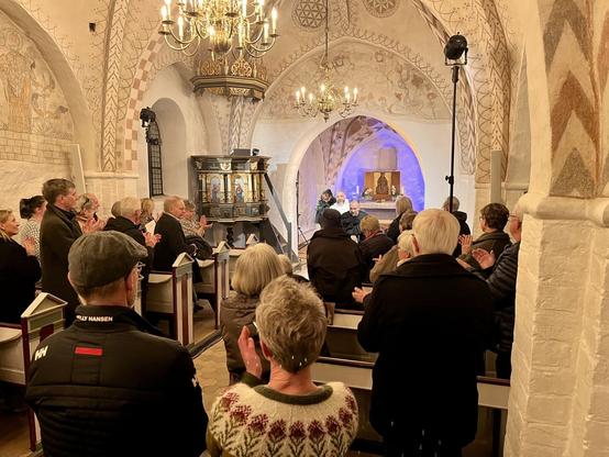 Vi er i en klassisk dansk kirke, en teaterforestilling er netop slut og publikum har rejst sig til stående applaus.

Kirkerummet er oplyst scenelyst.