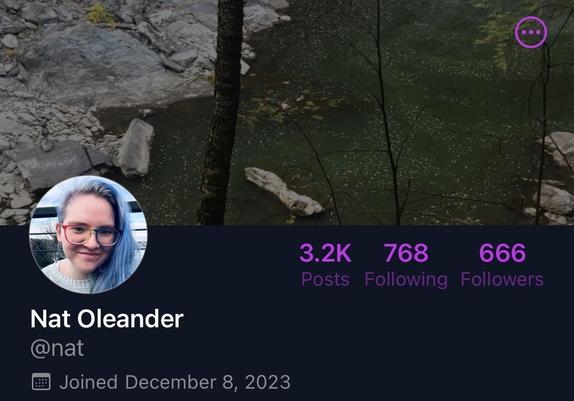 Screenshot of the top of Nat Oleander's profile, with 666 followers. Avatar is a white nonbinary person with blue hair and rainbow glasses who is smiling. Cover image is a greenish river with a rocky bank and a bare tree trunk in the middle of the photo.