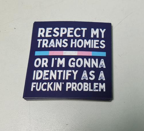 Square sticker, dark slate gray with white text and a ribbon in colors of the trans flag horizontally across the middle. It reads in bold capital typeface:
"Respect my trans homies -- or I'm gonna identify as a fuckin' problem"