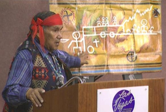 A photograph of a Native American man at a podium, pointing to an image from an ancient rock painting.