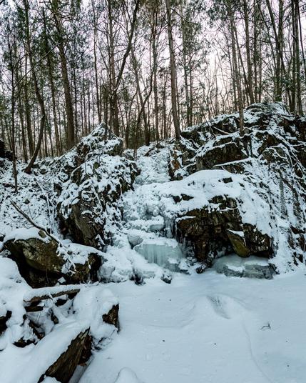 A frozen waterfall cutting through granite rocks in a wooded area. The area is covered in light snow