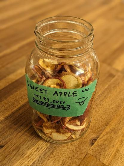 a clear glass canning jar sits on top of a wooden table. the jar is filled with many sliced, dehydrated apple chips sprinkled with cinnamon. the texture of the apple peel and the inner part of the apple are visible. a piece of green masking tape / painter's tape runs horizontally across the front of the jar, along with a cartoon smiling face (eyes closed, tongue barely sticking out, as if enjoying some delicious food) and the words "sweet apple cin.". the creation date is written on the tape - from October 2024.
