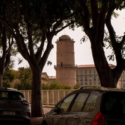 L'image comporte une grande tour ronde avec un sommet crénelé, peut-être une structure historique ou défensive. Il est encadré par des arbres au premier plan, créant un cadre naturel. Il y a des voitures garées visibles, indiquant un cadre de rue, et un bâtiment avec plusieurs fenêtres peut être vu en arrière-plan.