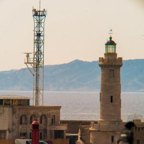 L'image comporte un phare et une tour de communication, près de l'eau. Il y a un bâtiment au premier plan, et des collines ou des montagnes sont visibles en arrière-plan à travers un plan d'eau. La scène donne une ambiance côtière ou portuaire.