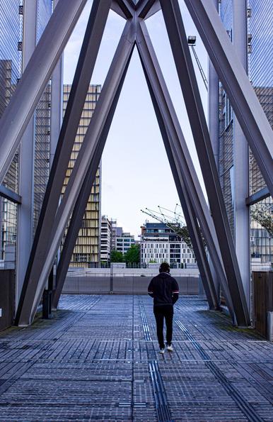 Une personne marche, on la voit de dos. Elle passe sous une structure métallique qui forme des grands compas.
