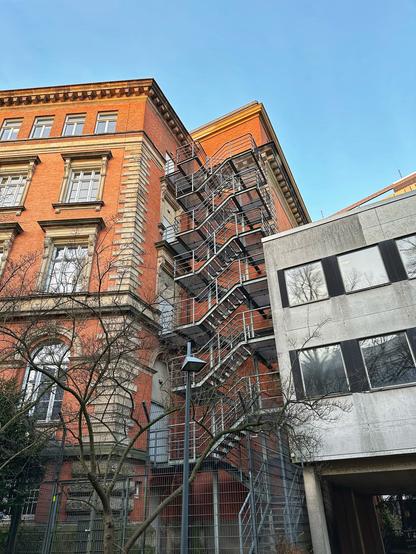 Ein rotes Backsteingebäude verfügt über eine markante externe Metalltreppe, die von Bäumen umgeben ist und sich von einer nahe gelegenen modernen Betonkonstruktion kontrastiert. Der Himmel ist oben klar.