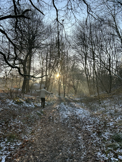 A frosty path through the woods with the sun filtering through the trees adding a golden glow to the snow. A man balances arms out as he walks down the path.