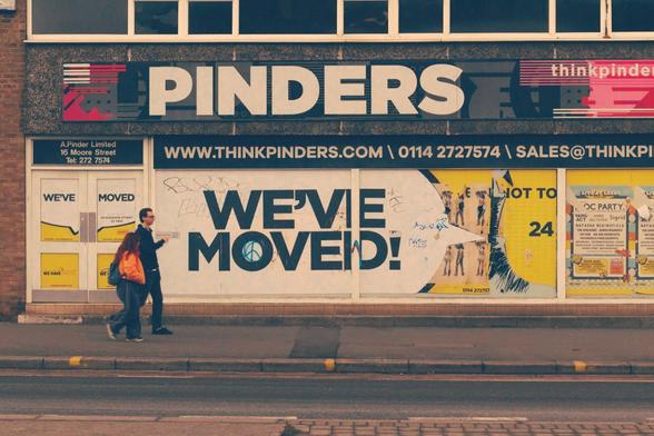 Two persons walking in front of a closed shop in sheffield. On the shop there's a banner saying we've moved and a small arrow pointing to the right. The two persons are moving in the same direction of the arrow giving the idea of following the suggestion