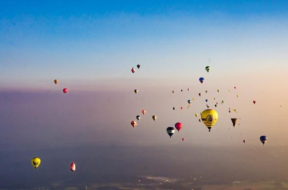 The image shows numerous colorful hot air balloons floating in the sky. They are set against a backdrop of a clear blue to warm gradient sky, possibly during sunrise or sunset. The ground is faintly visible at the bottom, suggesting an aerial view.