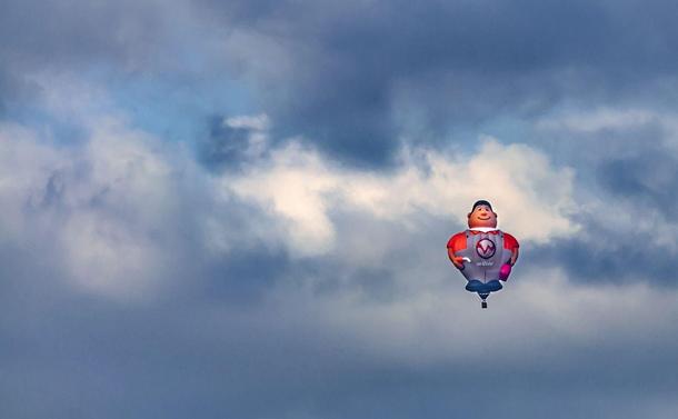 The image shows a hot air balloon designed to resemble a cartoonish character, possibly a superhero or pilot, with a helmet and a symbol on its chest. The balloon is set against a cloudy sky with patches of blue.