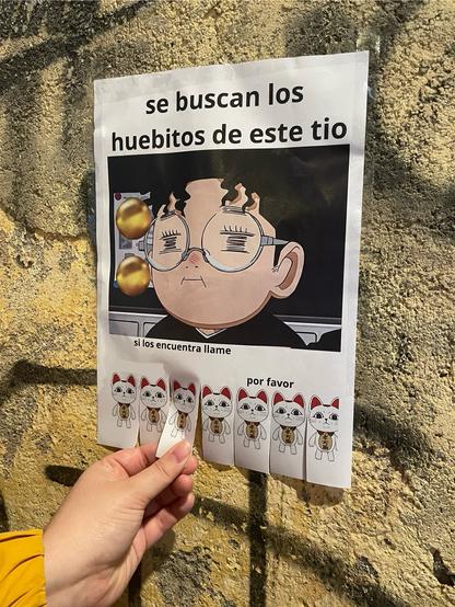 Cartel en un muro de la calle con tiras para arrancar. 

“se buscan los huebitos de este tío”

Captura del protagonista de Dandadan frunciendo la sonrisa y poniendo cara tonta. Dos bolas de oro a la izquierda de su cara.

“si los encuentra llame por favor”

En cada tira para arrancar hay un gato blanco de pie, es otro personaje de la serie. 

Yo estoy arrancando una de las tiras con mi mano.