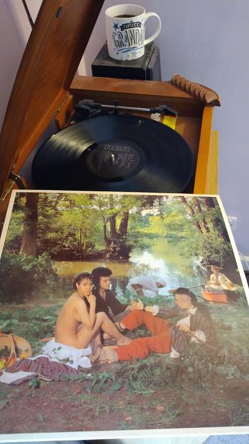 Record player with a 12" black vinyl record. In the foreground is an album cover showing two males and a naked female, looking at the camera, in a picnic group beside a tree lined pond.
Beyond this group, a woman is in the shallows, leaning forward and washing, and to the right a man sits rowing a small red dinghy.
Behind the player is a mug of coffee on a speaker and on the corner of the player is a delicious chocolate orange bar.
