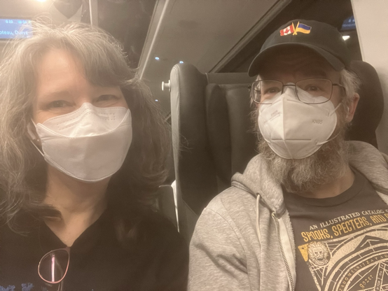 A woman with shoulder length greying hair wearing a white kn95 mask and a man with a ball cap with canada /ukraine flags, glasses, mask seating on a train
