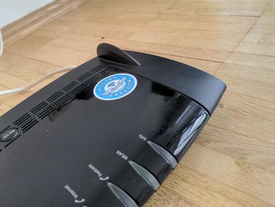 Black router with a round Router Freedom sticker on it. The router is laid down on a wooden floor.