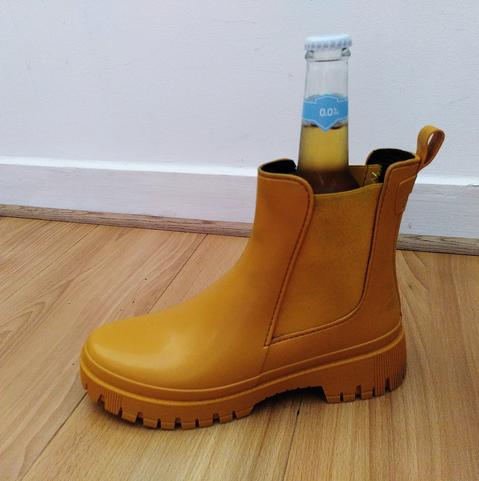 Bottine imperméable jaune posée sur du parquet stratifié beige, devant un mur blanc. Dans la bottine, le sommet d'une bouteille en verre à capsule avec un liquide jaune (du cidre) et une étiquette bleue claire portant la mention 0.0%