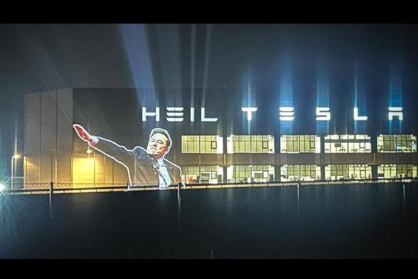 The factory has "Tesla" glowing across the top, but an image of Elon doing the Nazi salute and the word HEIL has been projected in front of it.