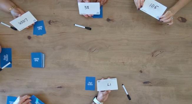 People playing the Magic Number card game at a table - just their hands and the cards, some cards flipped, numbers written, more close up