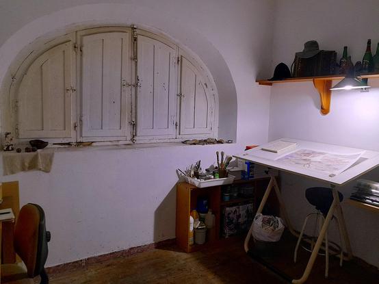 Una ventana en arco, cerrada con contraventanas de madera, al lado de una mesa de dibujo, iluminada por un foco de luz enganchado a una estantería de madera. llena de objetos entrañables.