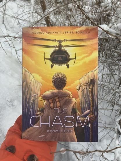 A paperback of The Chasm being held outside above snow. The cover features a bright yellow-orange sunset behind a hovering black helicopter. A woman in arctic clothing and a walking stick defiantly faces off against the helicopter.