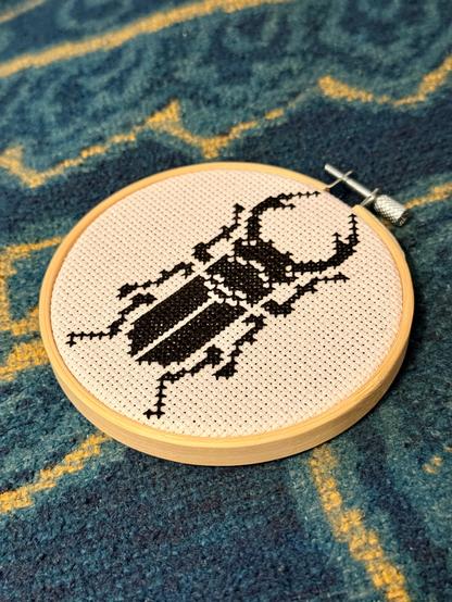 Stickrahmen auf eine gelb-petrol-farbenen Decke. Der Aida-Stoff im Rahmen wurde mit einem etwa 10cm großen Motiv eines männlichen Hirschkäfers in schwarz per Kreuzstich bestickt.