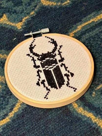 Stickrahmen auf eine gelb-petrol-farbenen Decke. Der Aida-Stoff im Rahmen wurde mit einem etwa 10cm großen Motiv eines männlichen Hirschkäfers in schwarz per Kreuzstich bestickt.