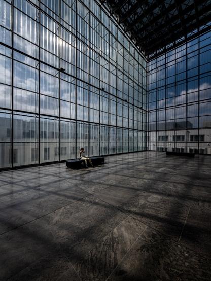 The photo shows a large, light-filled hall in the Museum of Fine Arts in Leipzig. The walls and ceiling consist of a grid-like steel and glass structure, offering a view of the sky and surrounding buildings. The glass surfaces reflect the incoming light, creating a pattern of shadows and lines on the gray stone floor. In the right foreground, a single person sits on a dark bench, their white top and golden pants subtly contrasting with the minimalist environment. The hall appears spacious and modern.