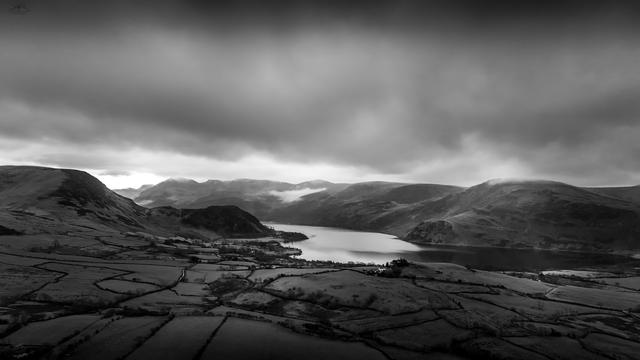 Monochrome aerial shot over fields towards lake in background surrounded by mountains
