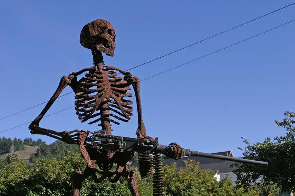 In the background are clear blue sky, trees, hills and a partial view of a house.

In the foreground is an 8-foot skeleton wielding a .50-caliber M2HB machine gun, "from the hip." There is a short belt in the MG.