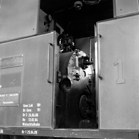 The black-and-white photo provides a glimpse into the heart of a steam locomotive. The viewer looks directly at a large, cylindrical boiler surrounded by numerous shiny metal parts. Various levers, wheels, and gauges can be seen, which were important for controlling the machine. In the background, the contours of other machine elements can be guessed. The overall image conveys an impression of power, precision, and the era of steam technology.