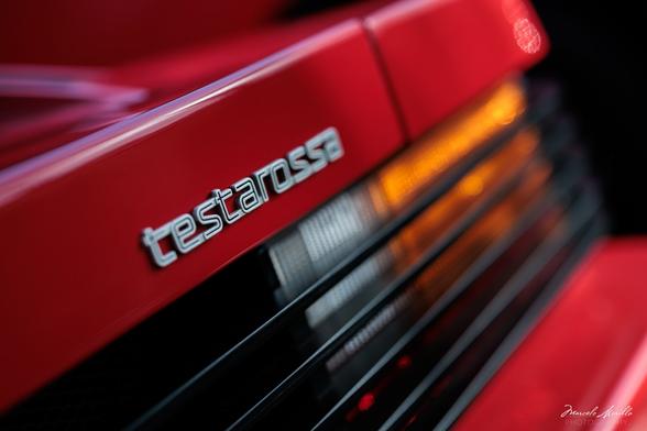 Close-up image of a Ferrari Testarossa badge on a red car, with the focus on the silver “Testarossa” emblem and blurred rear grille and indicator lights in the background.