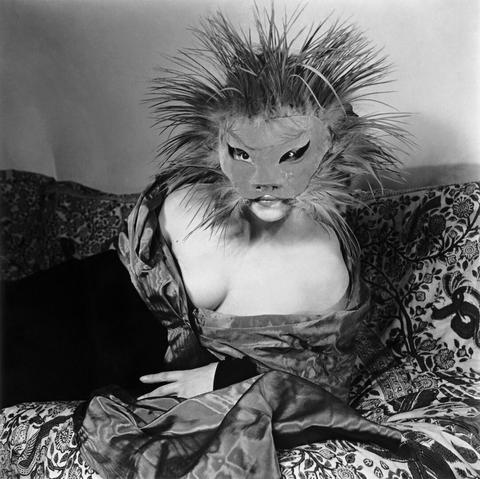 A back and white photo of an elegant woman reclining on a brocade upholstered couch. She wears an off the shoulder gown of black velvet trimmed with taffeta and a large feline mask bristling with feathers that resemble a lion’s mane
