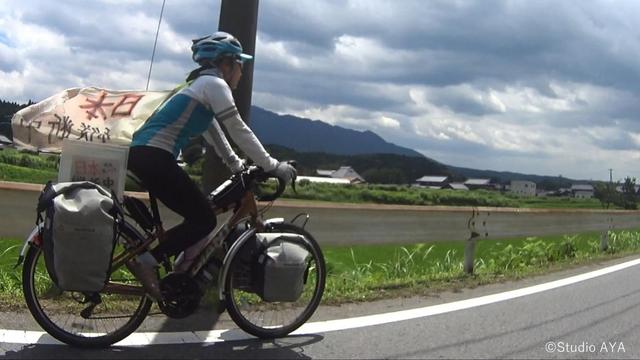 Regisseurin Ayako Imamura fährt auf ihrem Fahrrad mit Gepäcktaschen vorne und hinten auf einer asphaltierten Straße vor wolkenverhangener Kulisse. Im Hintergrund ein Hügel.
