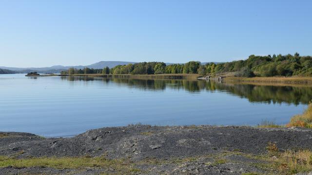 A photo of a bay. The foreground is rocky and a peninsula to the right has wetland along it's shores and forests on it. There are hills in the distance. The sky is clear and blue.