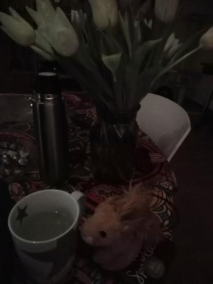 Puschel sitzt neben weißer Tasse mit nem grauen Stern drauf, auf net bunten Tischdecke. Im Hintergrund stehen Tulpen in weiß und lila. Das Bild ist spärlich beleuchtet.