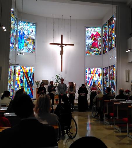 Gottesdienstgemeinde in der Familienkirche, im Hintergrund hängt ein großes Holzkreuz von der Decke; links und rechts sind Buntglasfenster zu sehen; vor dem Altar stehen sechs Personen, zwei davon Borromäerinnenschwestern, die Gaben zum Altar gebracht haben und Fürbitten vortragen (© privat)