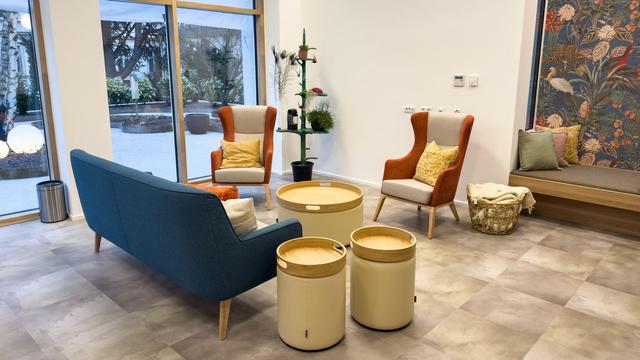 Audenthaltsraum im neuen Tageshospiz mit Sofa, Tonnentischen und zwei Armsesseln; im Eck sind Pflanzen, links geht es auf die Terasse hinaus; rechts eine Sitznische mit Pölstern und einem Wandbild mit blauem Hintergrund mit Vögeln und Seepflanzen (© CS)