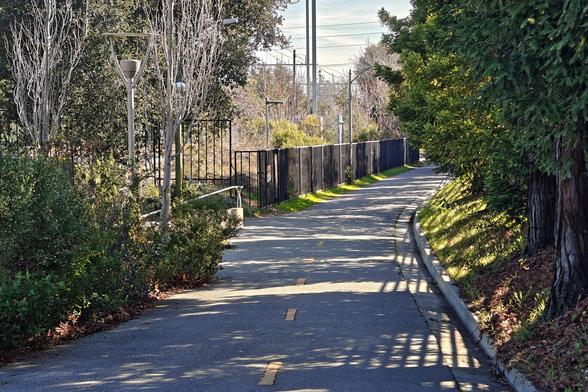 An yellow-striped asphalt path curves away and off to the right. Trees line the right side of the path, and a security fence edges the path on the left.