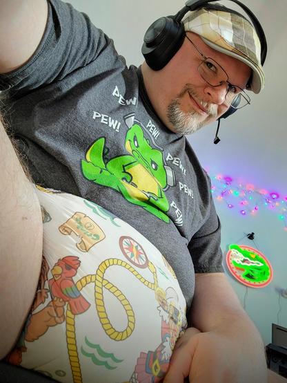 A middle-aged man, wearing a t-shirt, cap, headset, and obvious adult diaper, grins at the camera knowingly.