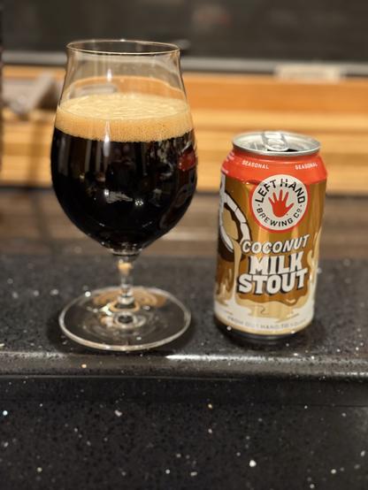 A tall stout snifter of dark beer next to a gold tone can with a red top and a coconut pouring coconut milk